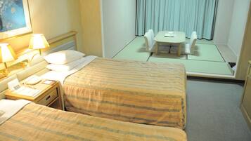 Une chambre d’hôtel avec deux lits, une table de chevet avec une lampe, une petite table avec une chaise, et une vue sur un balcon avec des rideaux.