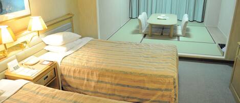 A hotel room with two beds, a bedside table with a lamp, a small table with a chair, and a view of a balcony with curtains.