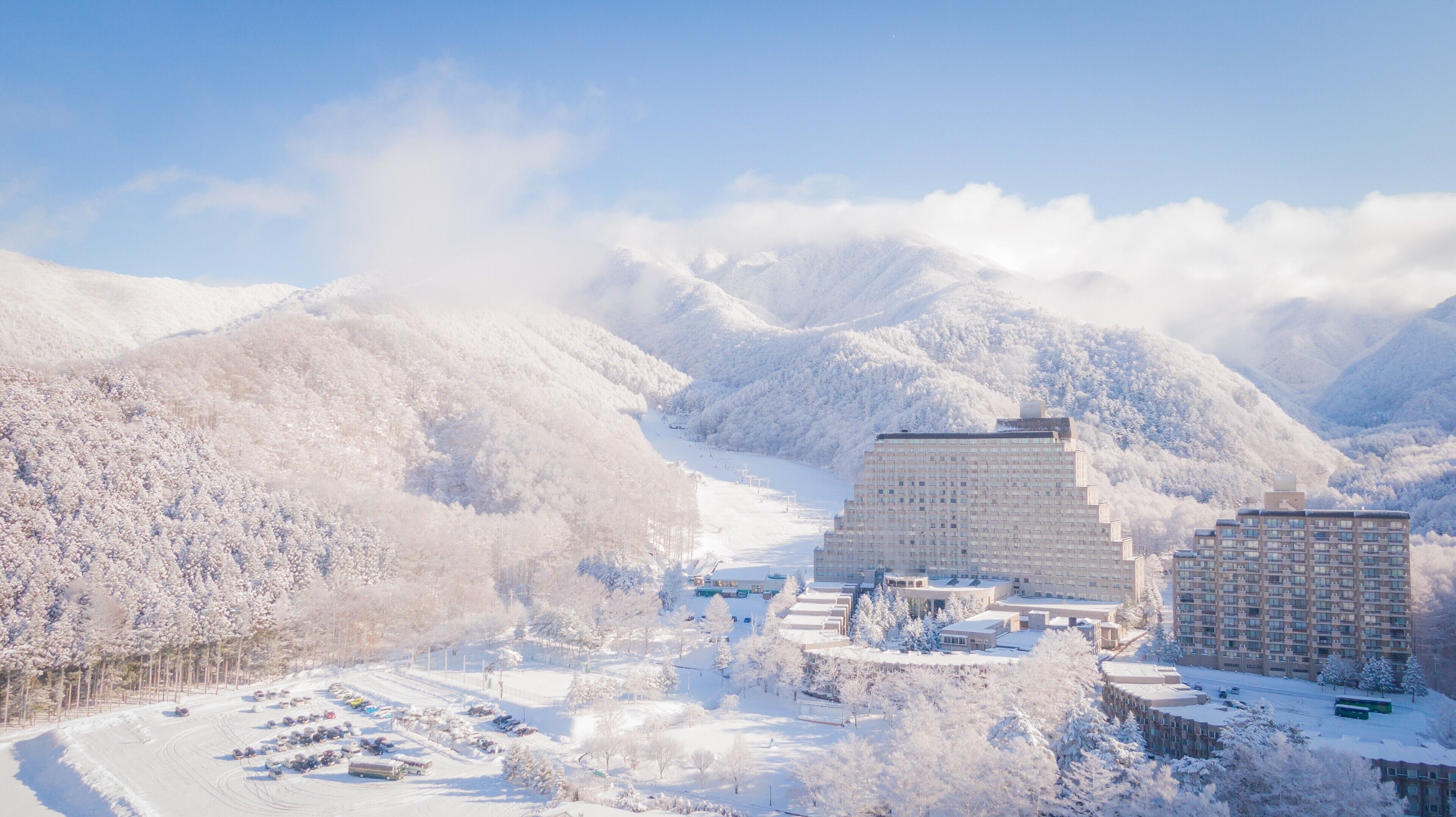 A snow-covered mountainous area with a large building complex, including a multi-story hotel and smaller structures, surrounded by trees and a parking area.