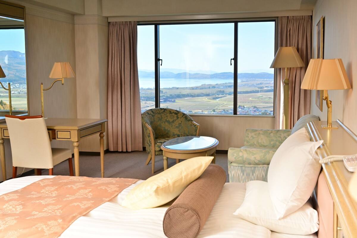 A hotel room with a bed, a desk, a chair, a small table, and a view of the landscape through large windows.
