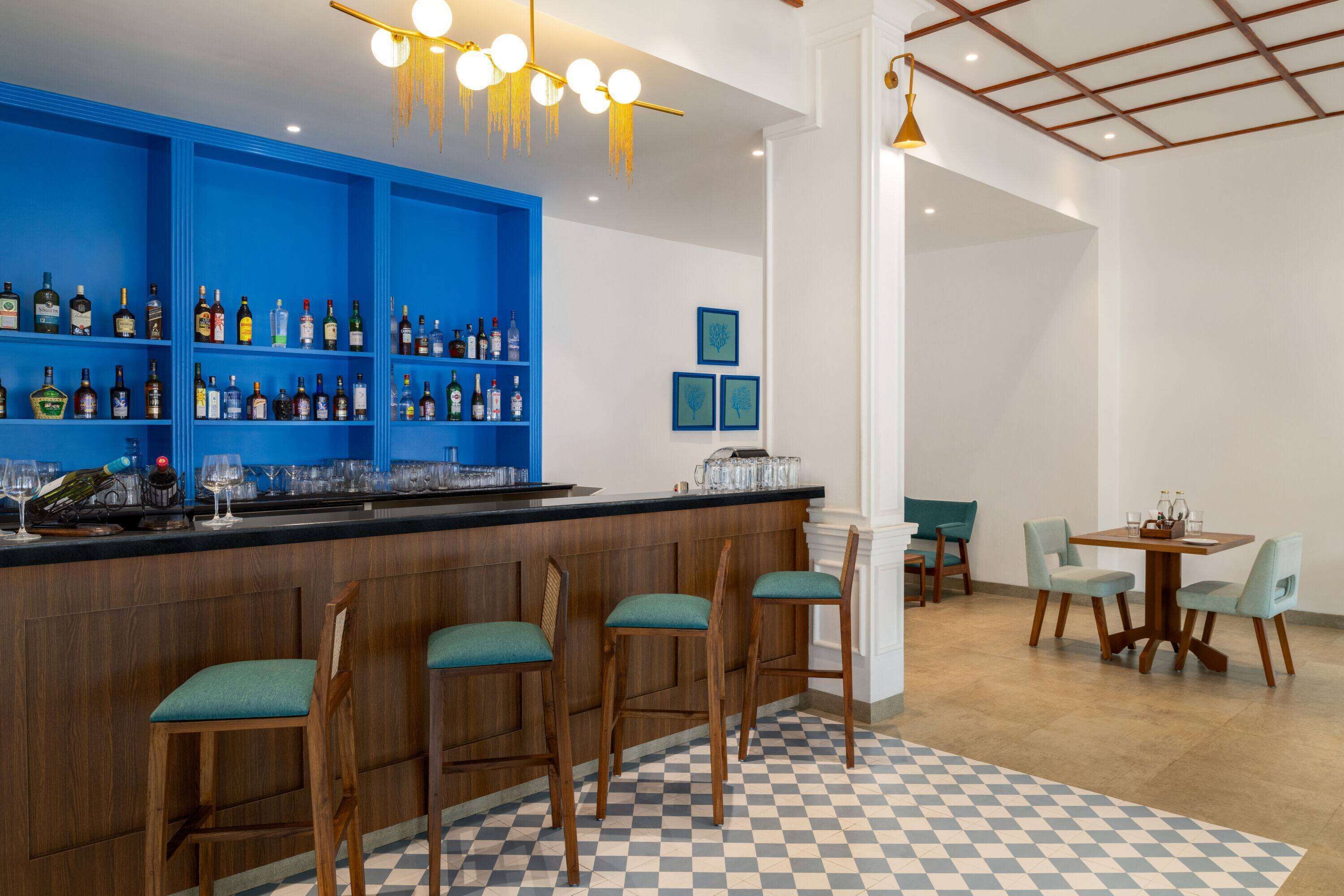 A bar area with blue shelves, wooden bar counter, and bar stools. There is a checkered floor and a dining area with a table and chairs.