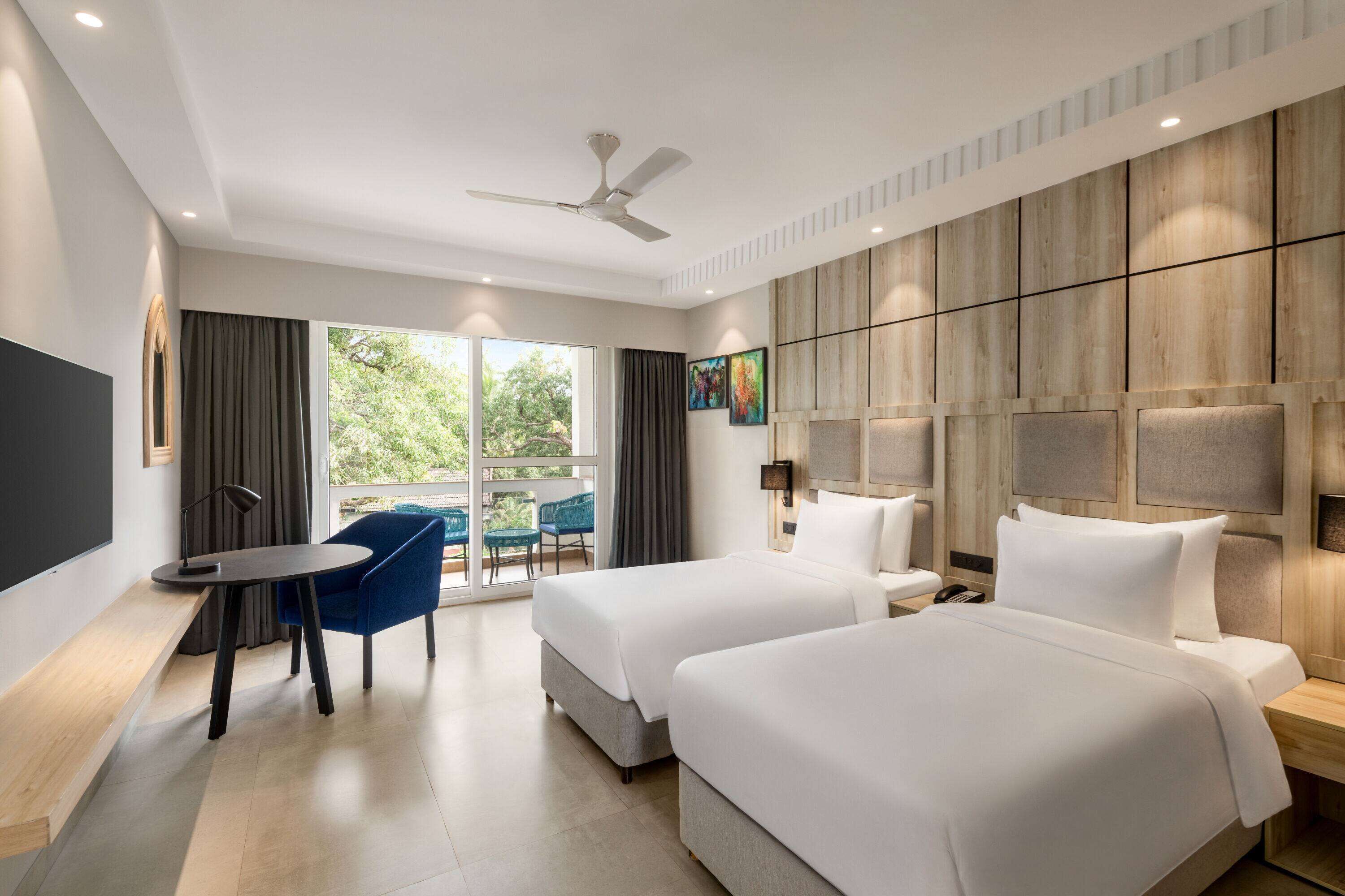 A hotel room with two beds, a desk, a chair, and a large window with curtains.