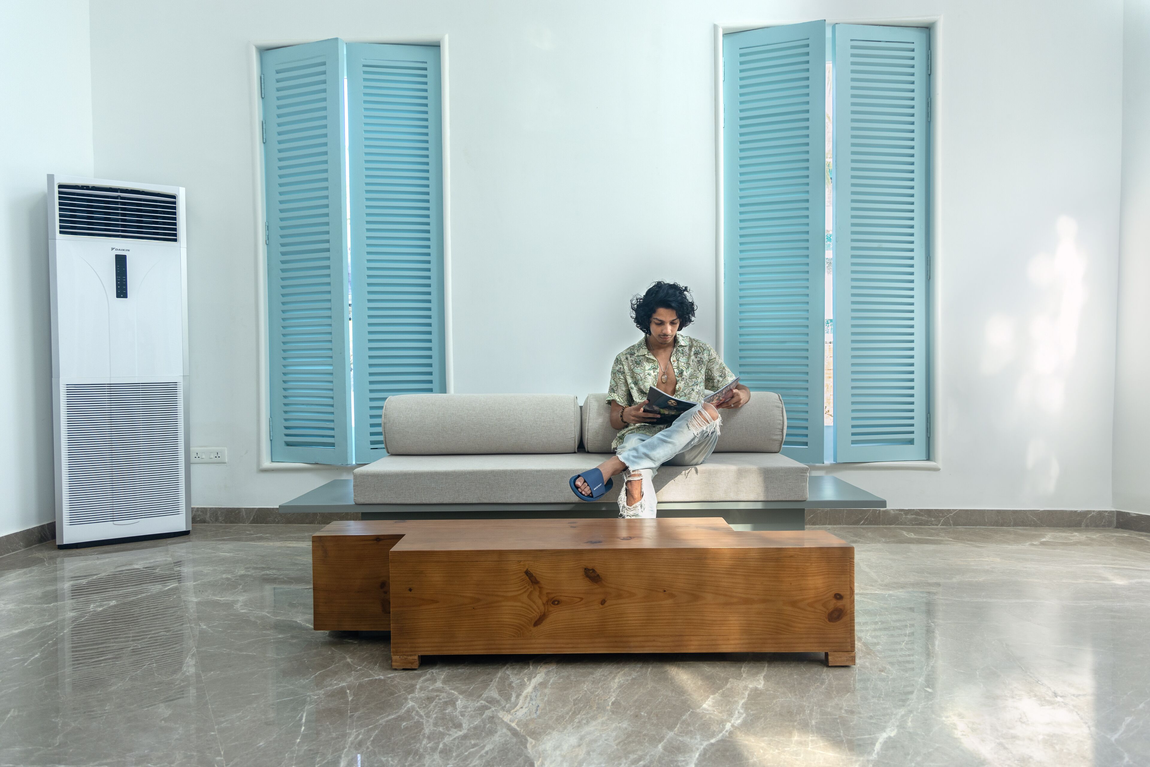 A person sitting on a sofa in a room with a large air conditioner and blue shutters on the windows.