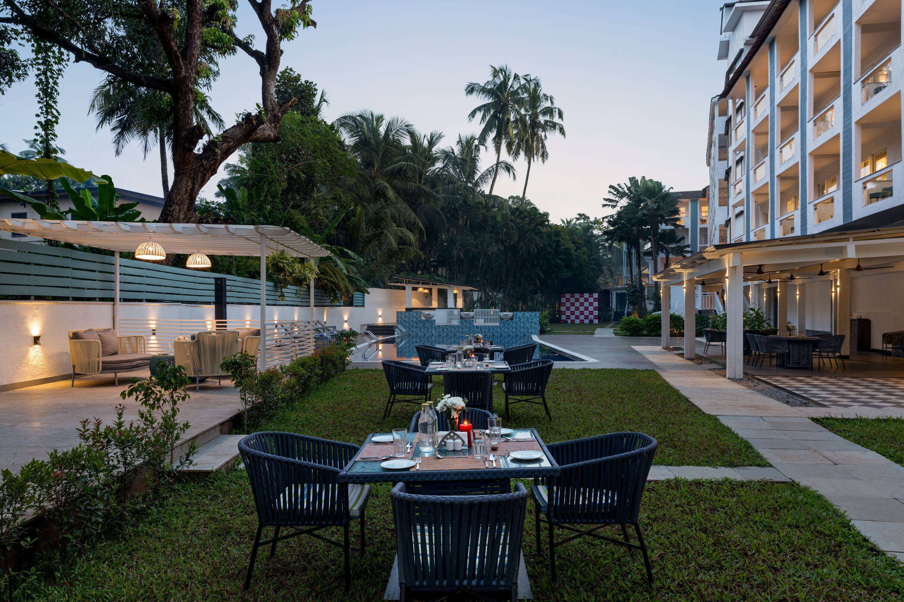 Un área de comedor al aire libre con mesas y sillas, rodeada de follaje y palmeras.