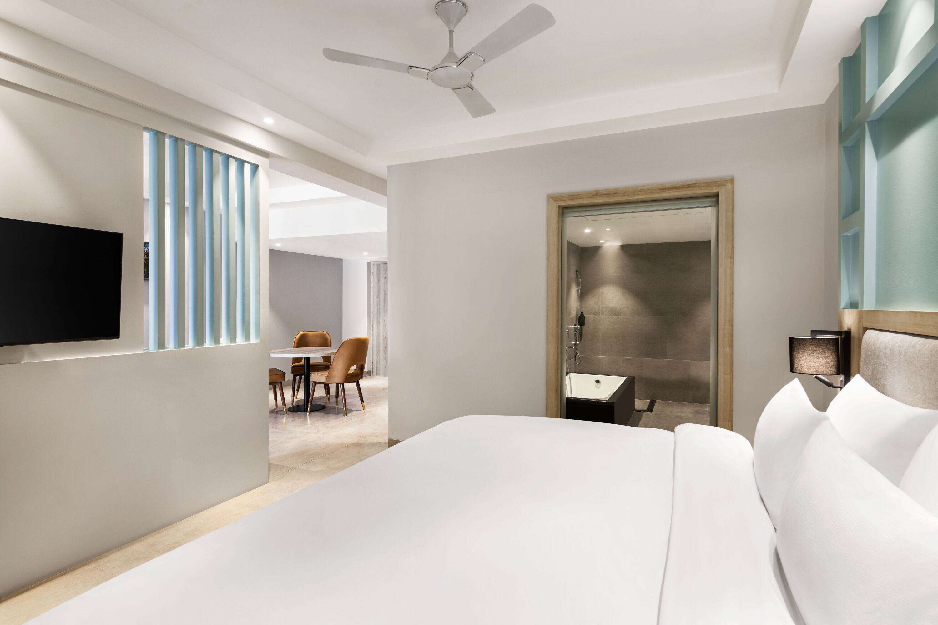 A hotel room with a bed, a TV mounted on the wall, a ceiling fan, a bathroom with a mirror, and a dining area with chairs.