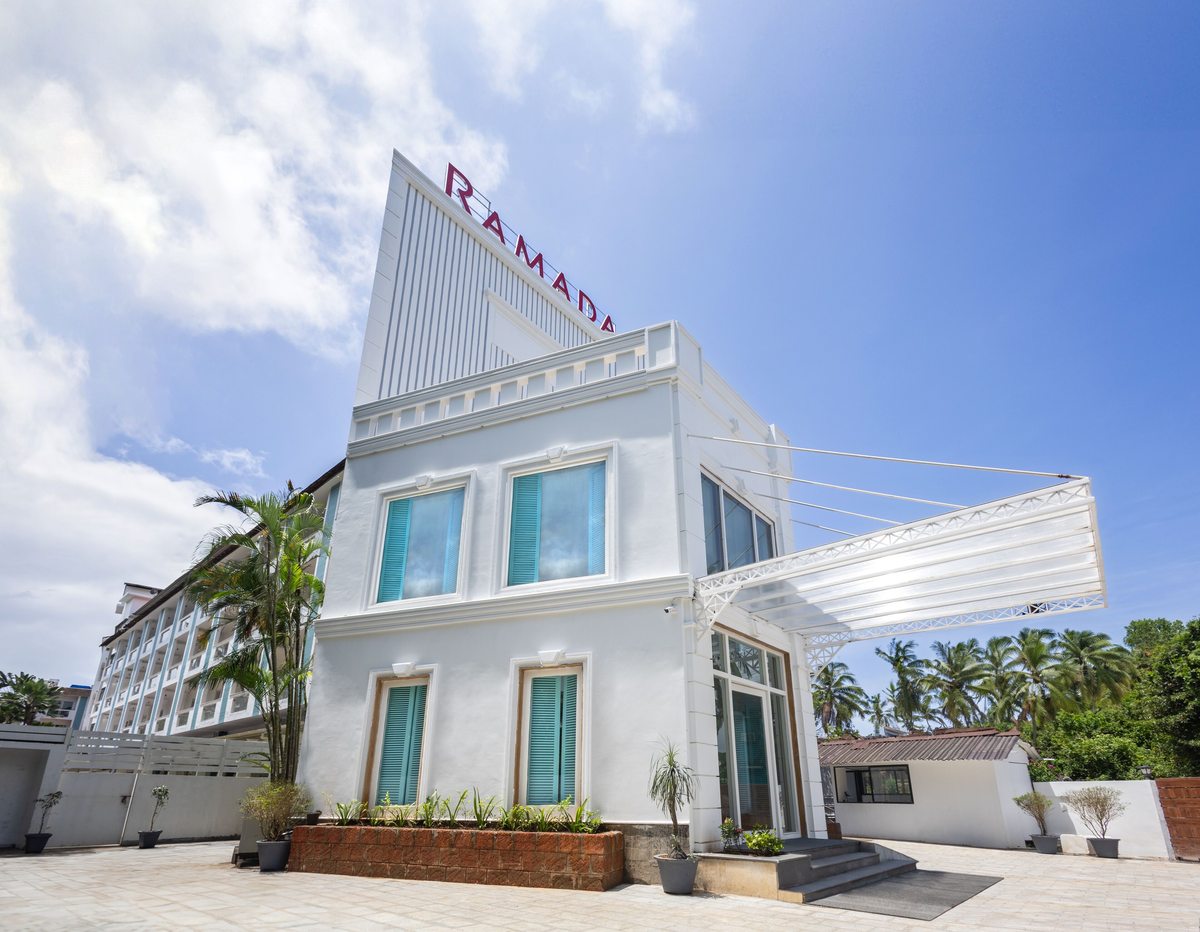 Un edificio moderno con un llamativo letrero 'RAMAD', ventanas amplias y un área exterior cubierta con plantas en macetas.