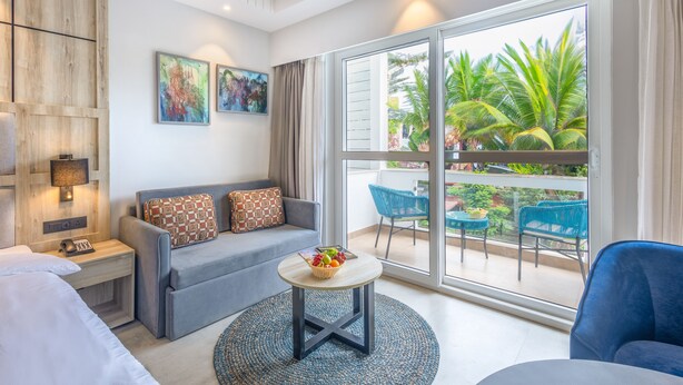 A modern hotel room with a grey sofa, a small round table with fruit, a balcony with blue chairs, and a large window with a view of palm trees. - Ramada by Wyndham Goa Arpora (Arpora)