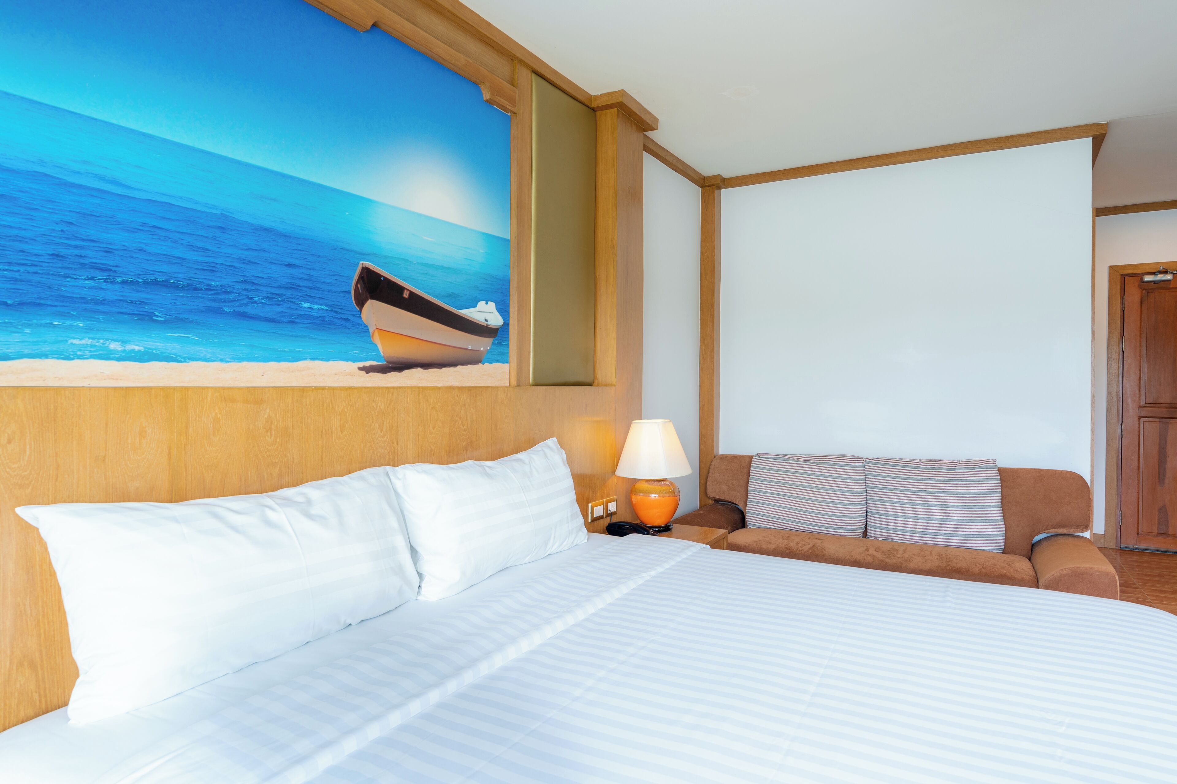 A hotel room with a bed, a nightstand, a lamp, a sofa, and a painting of a boat on the wall.