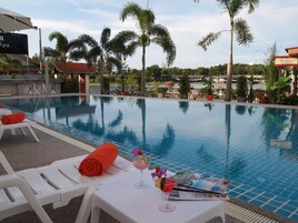 Un área de piscina con sillones reclinables, una mesa con una bebida y una revista, y palmeras al fondo.