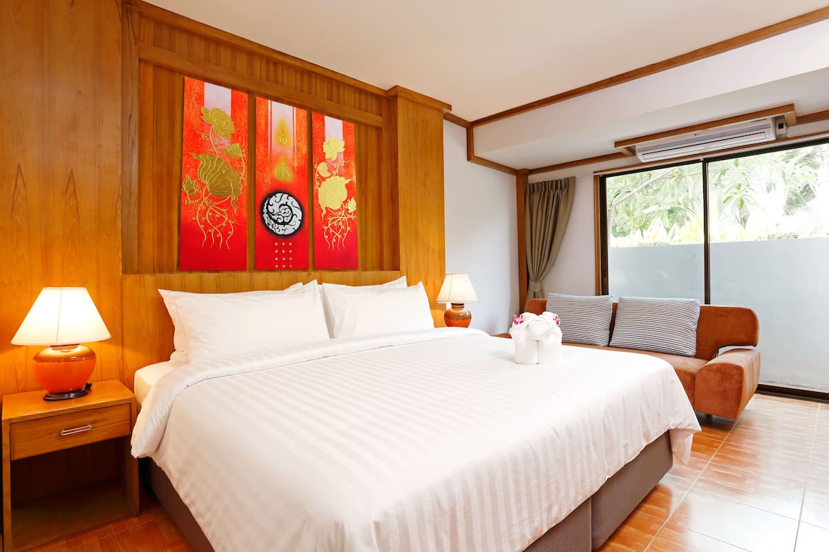 A hotel room with a large bed, two bedside tables with lamps, a sofa, a large window with a view of greenery, and a wall with traditional artwork.