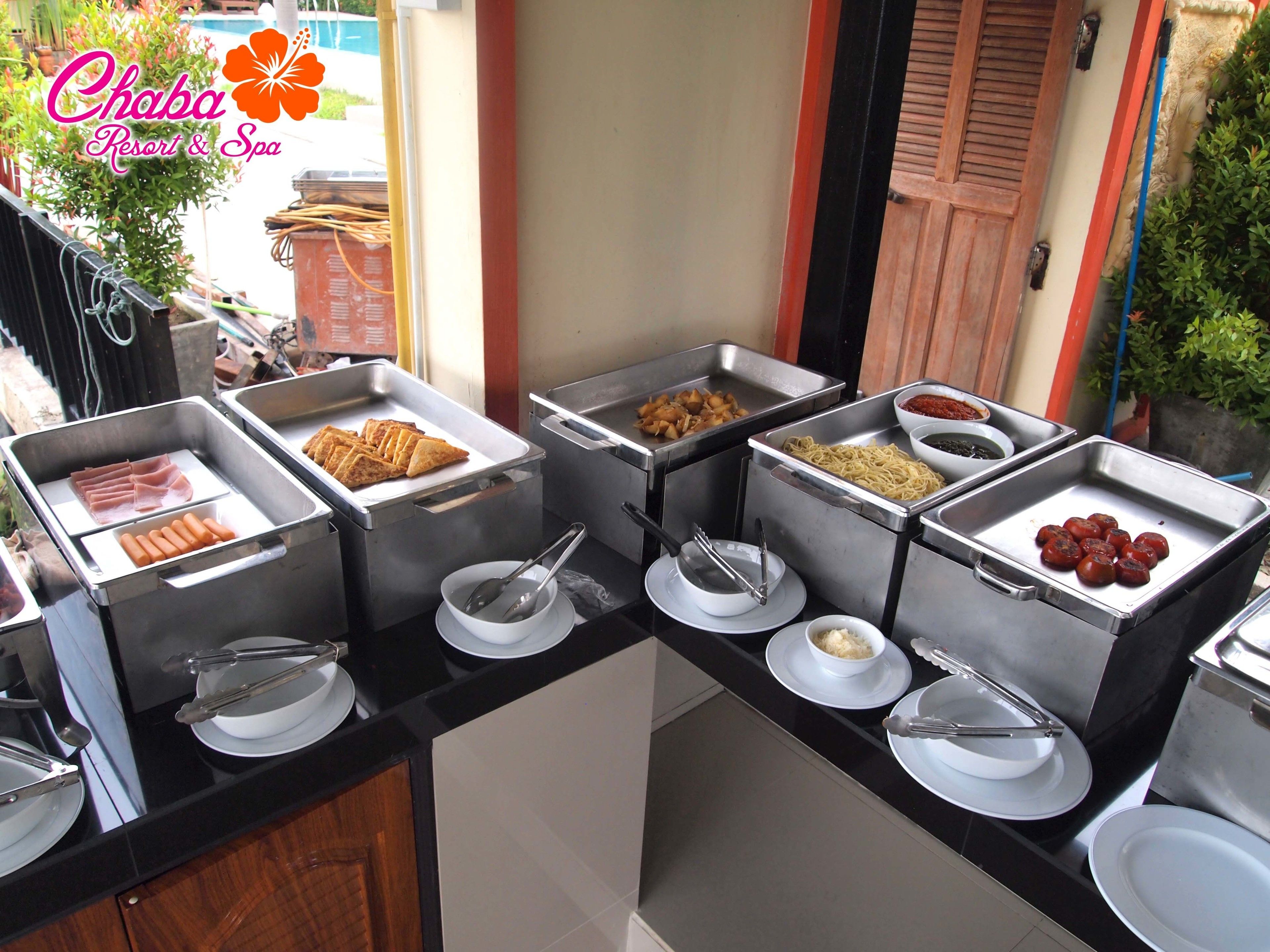 A buffet setup with various food items in metal trays, including hot dogs, samosas, and other dishes, with serving utensils and plates.