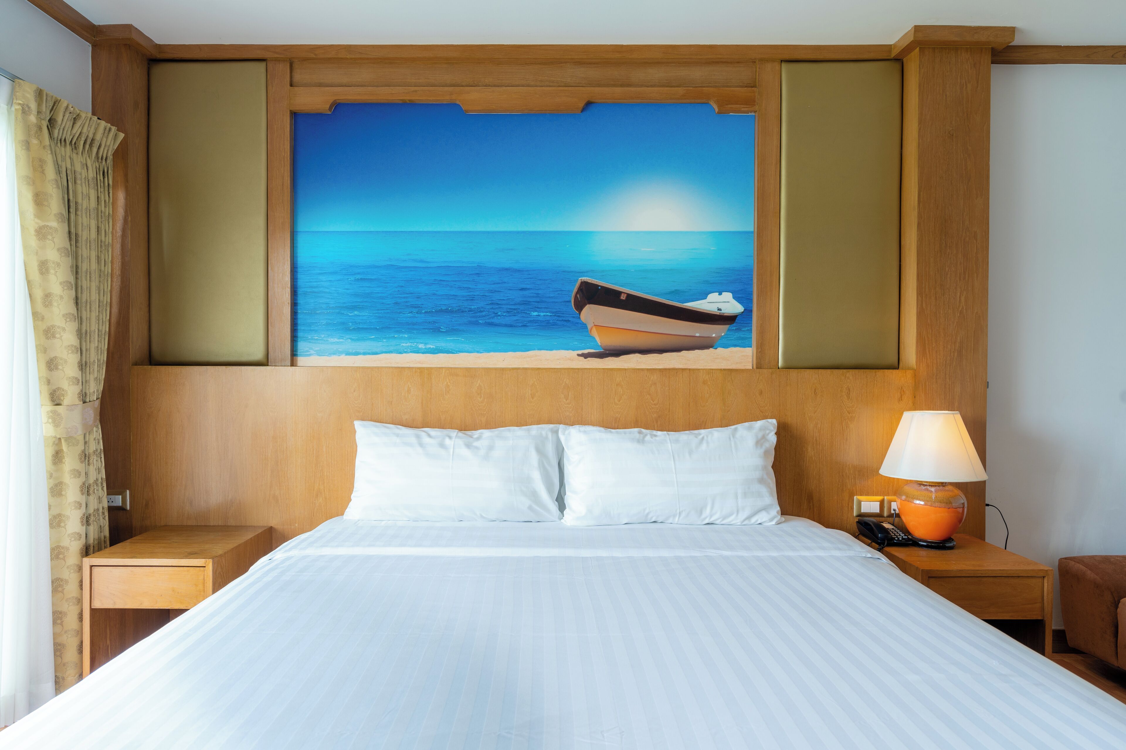 Ein Hotelzimmer mit Bett, Nachttisch mit Lampe, einem Strandbild und einem Holzkopfstück.