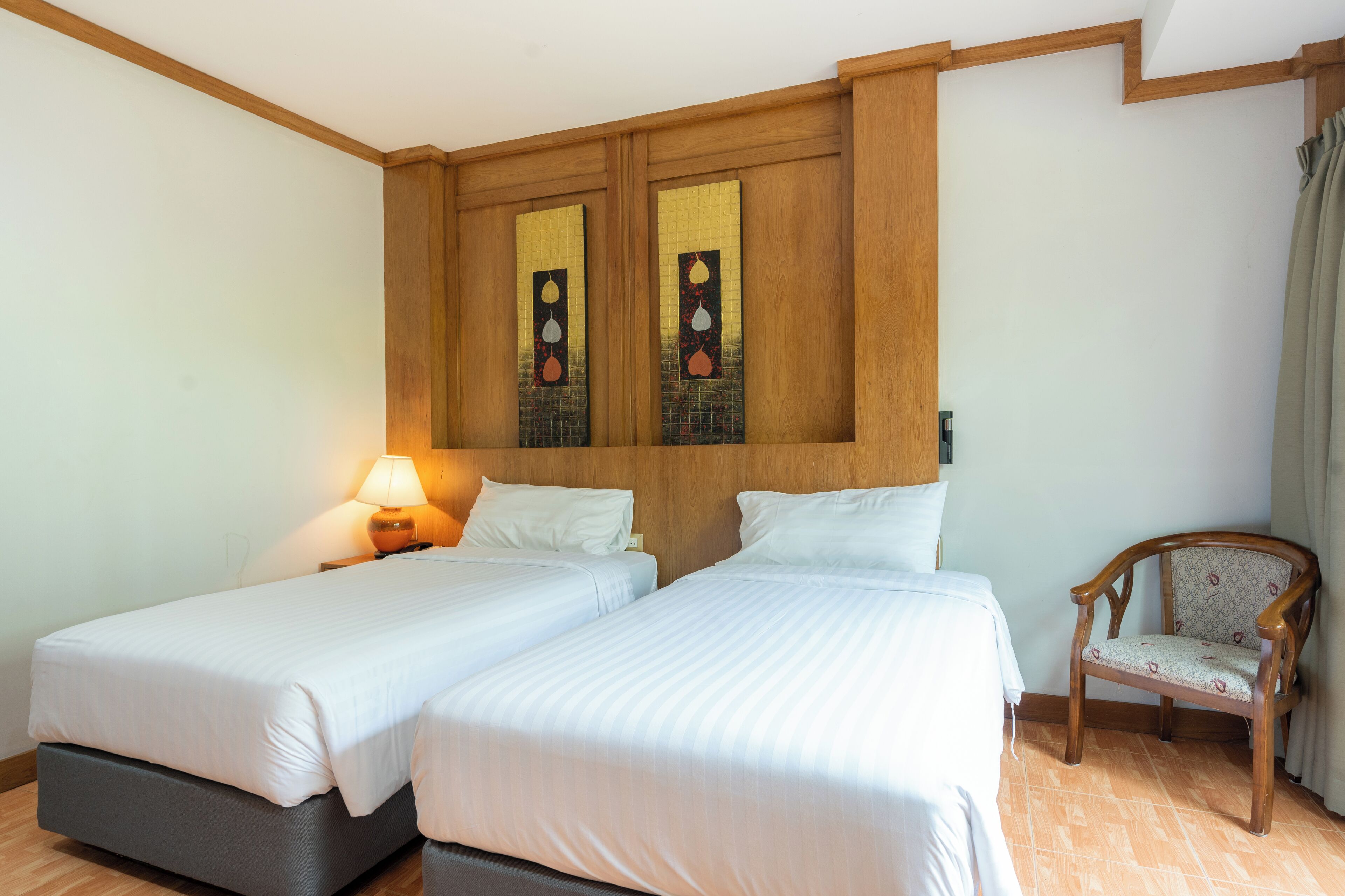 A hotel room with two beds, a wooden headboard with artwork, a chair, and a lamp.