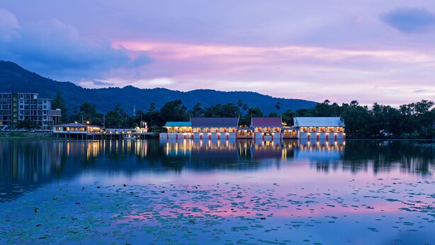 Hotel junto a un lago, con el reflejo de edificios en el agua, rodeado de montañas y un cielo al atardecer.