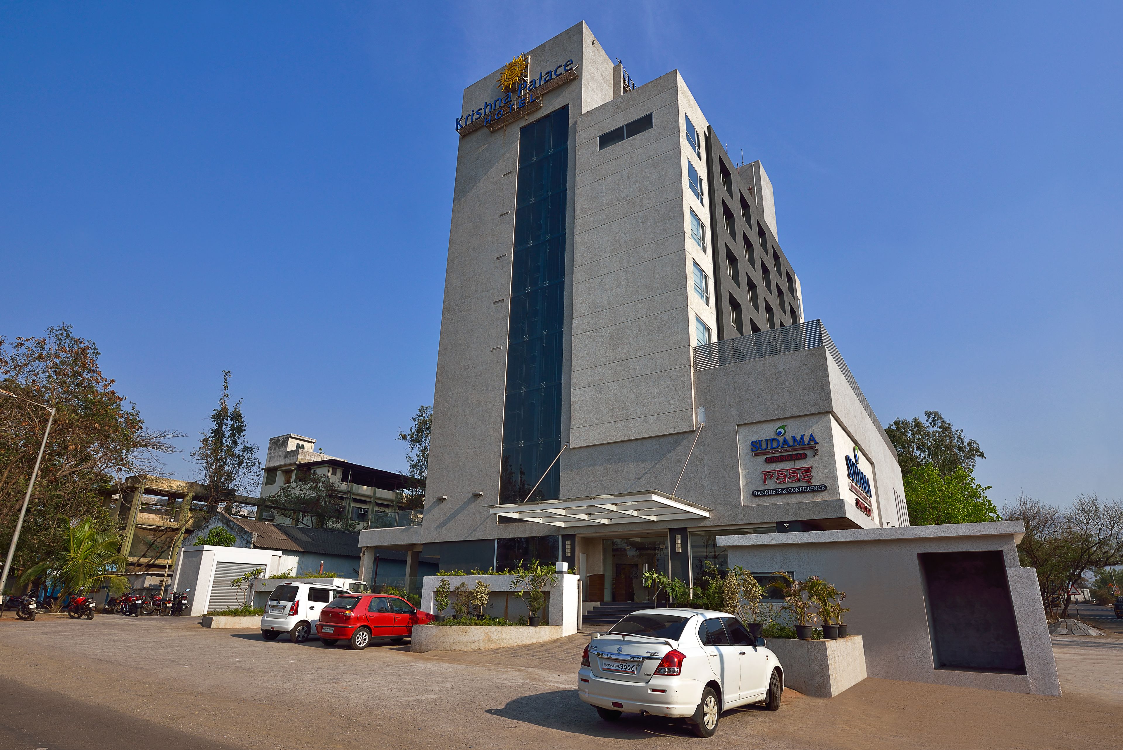 A modern hotel with a prominent entrance, a parking area with several cars, and a clear blue sky.
