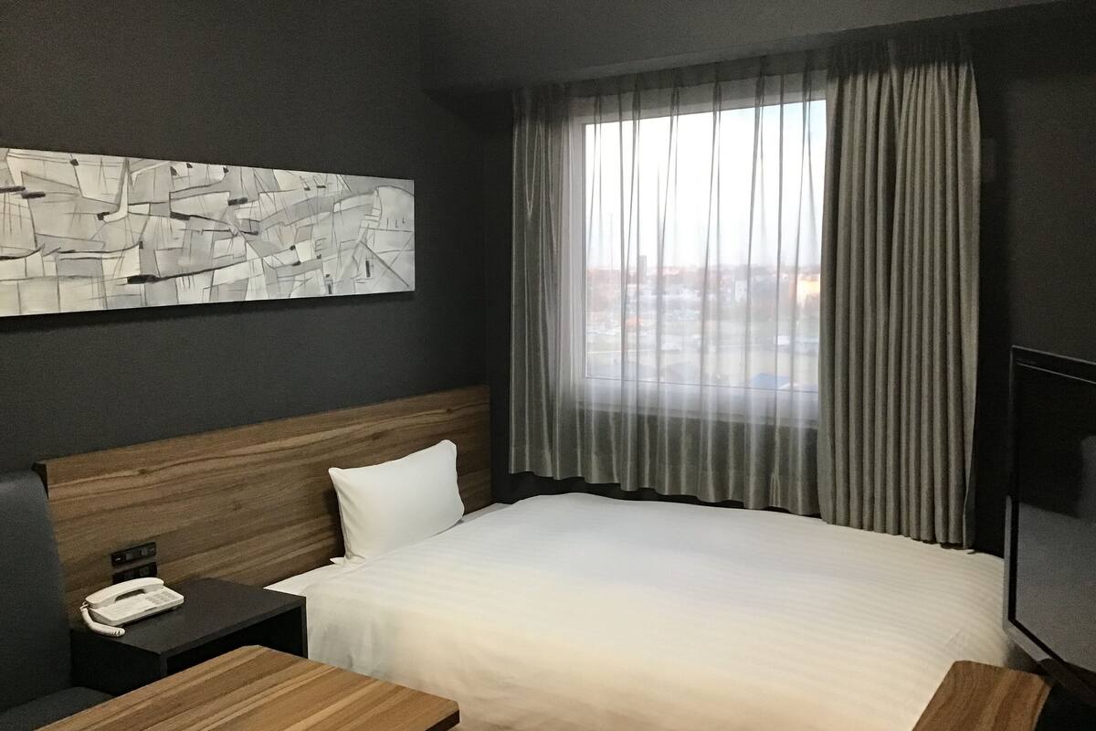 A hotel room with a large bed, a wooden headboard, a bedside table with a phone, a TV, and a window with curtains.