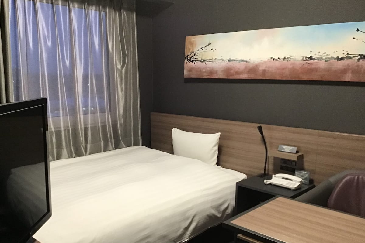 A hotel room with a bed, a bedside table with a phone, a small desk, and a painting on the wall.
