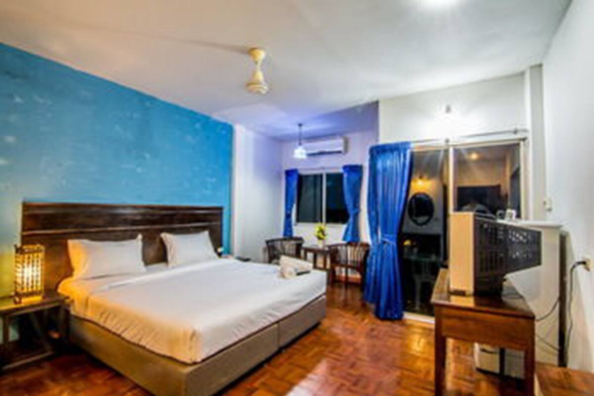 Quarto de hotel com uma cama grande, uma escrivaninha com computador, uma janela com cortinas e um ventilador de teto.