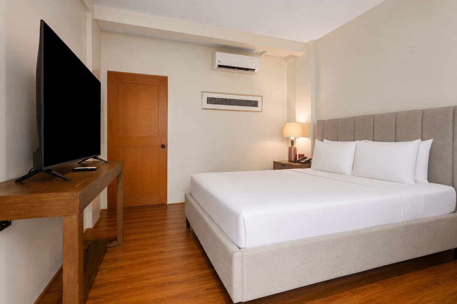 A hotel room with a bed, a desk with a television, a bedside lamp, and an air conditioning unit.