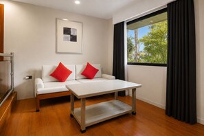 Una habitación moderna con un sofá blanco, dos cojines rojos, una mesa de madera y una ventana con cortinas oscuras.