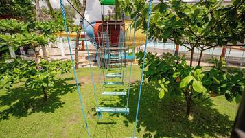 A rope swing set in a grassy area with trees and a playground structure in the background.