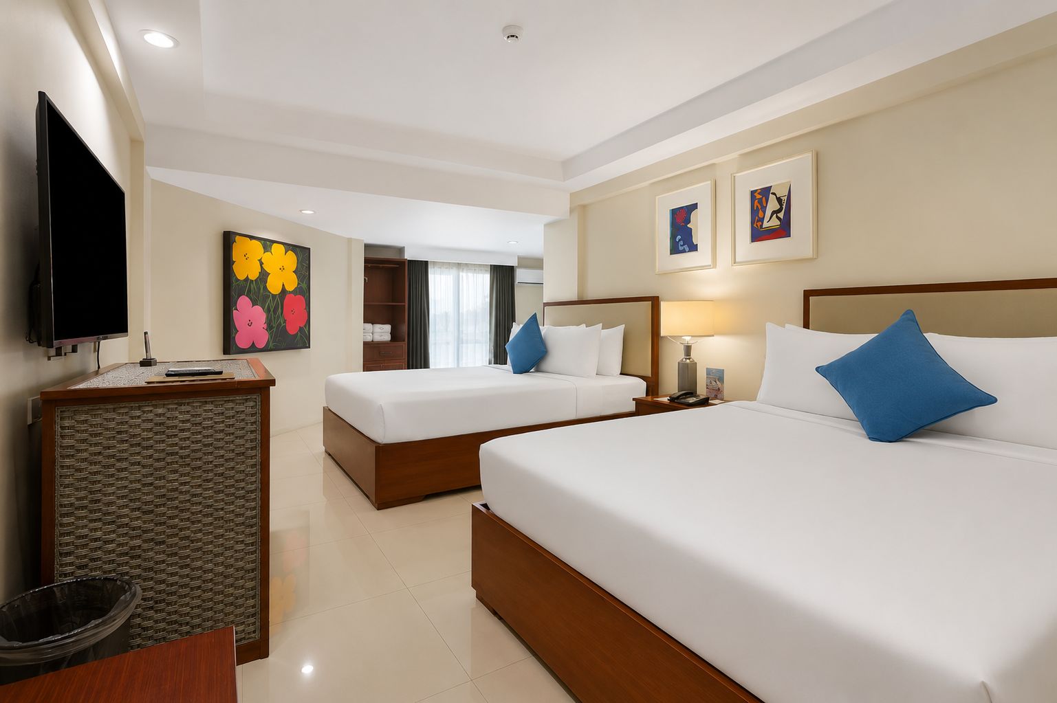 A hotel room with two beds, a television, artwork on the walls, and a desk with a lamp.