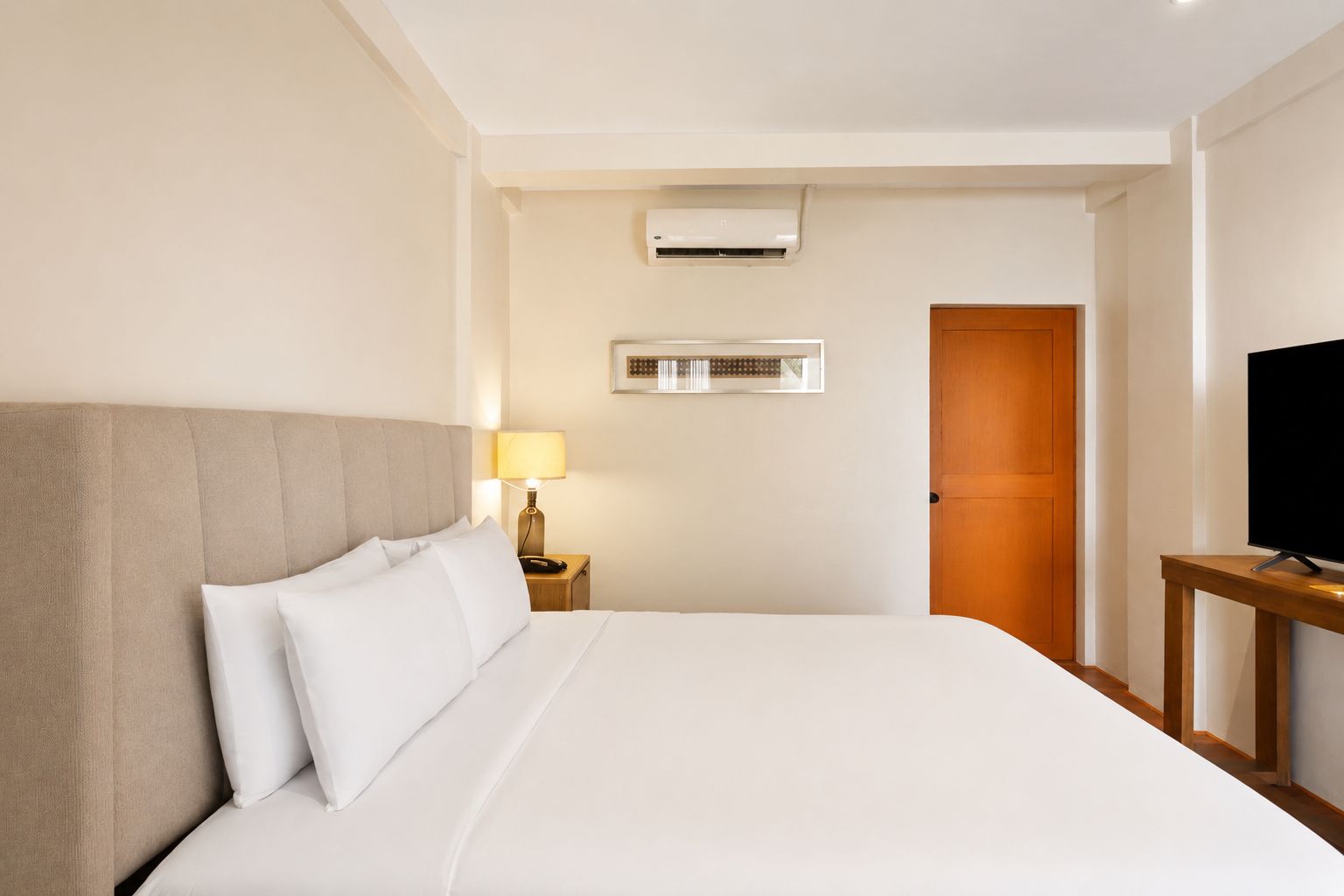 A hotel room with a bed, a bedside lamp, an air conditioning unit, a television on a stand, and a wooden door.