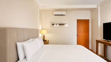 A hotel room with a bed, a bedside lamp, an air conditioning unit, a television on a stand, and a wooden door.
