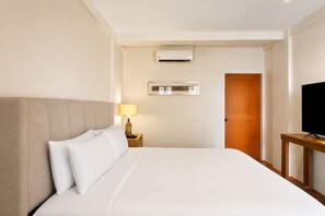 Una habitación de hotel con cama, lámpara de noche, equipo de aire acondicionado, televisor sobre un mueble y puerta de madera.