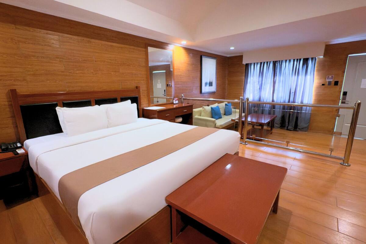 A hotel room with a large bed, wooden floor, a desk, a chair, a sofa, a mirror, and a window with curtains.