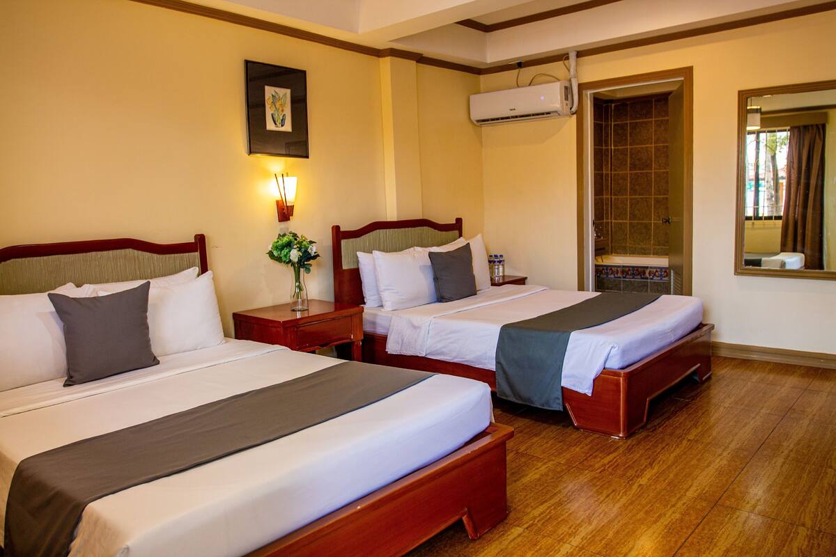 A hotel room with three beds, a wooden floor, a wall-mounted air conditioner, and a framed picture.