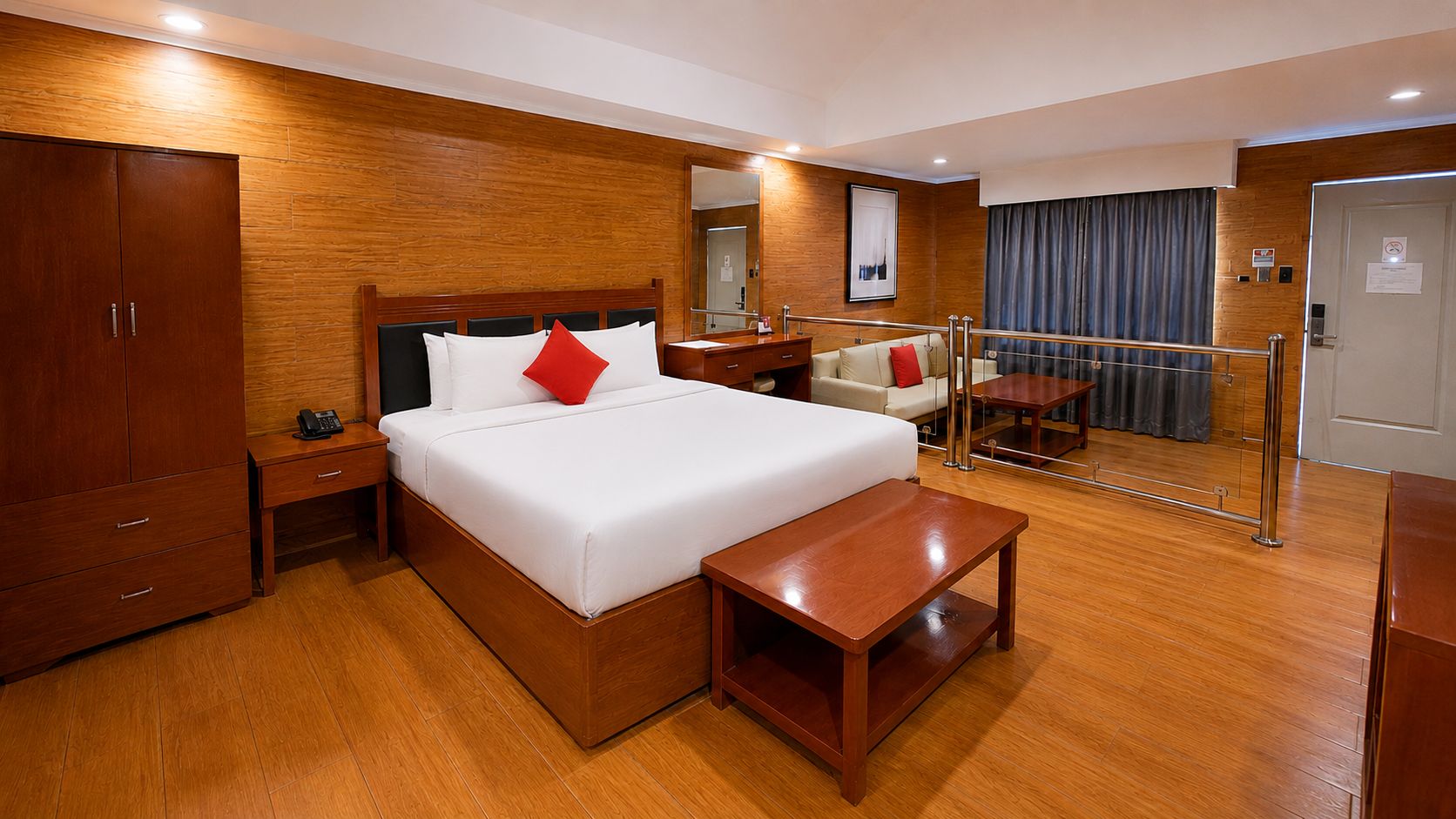 Une chambre d’hôtel avec un plancher en bois, un lit avec une literie blanche et un oreiller rouge, une table de chevet, une armoire en bois, un miroir, un coin salon avec un canapé et une petite table, et une porte avec une pancarte.