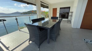 Un balcón con mesa de vidrio y sillas de mimbre, con vista a un cuerpo de agua y colinas.