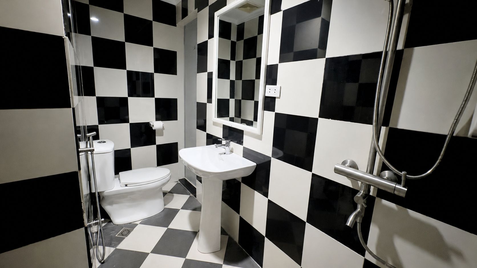 Une salle de bain avec un carrelage noir et blanc à motifs de damier, un lavabo blanc, des toilettes et une douche équipée d’un pommeau de douche à main.