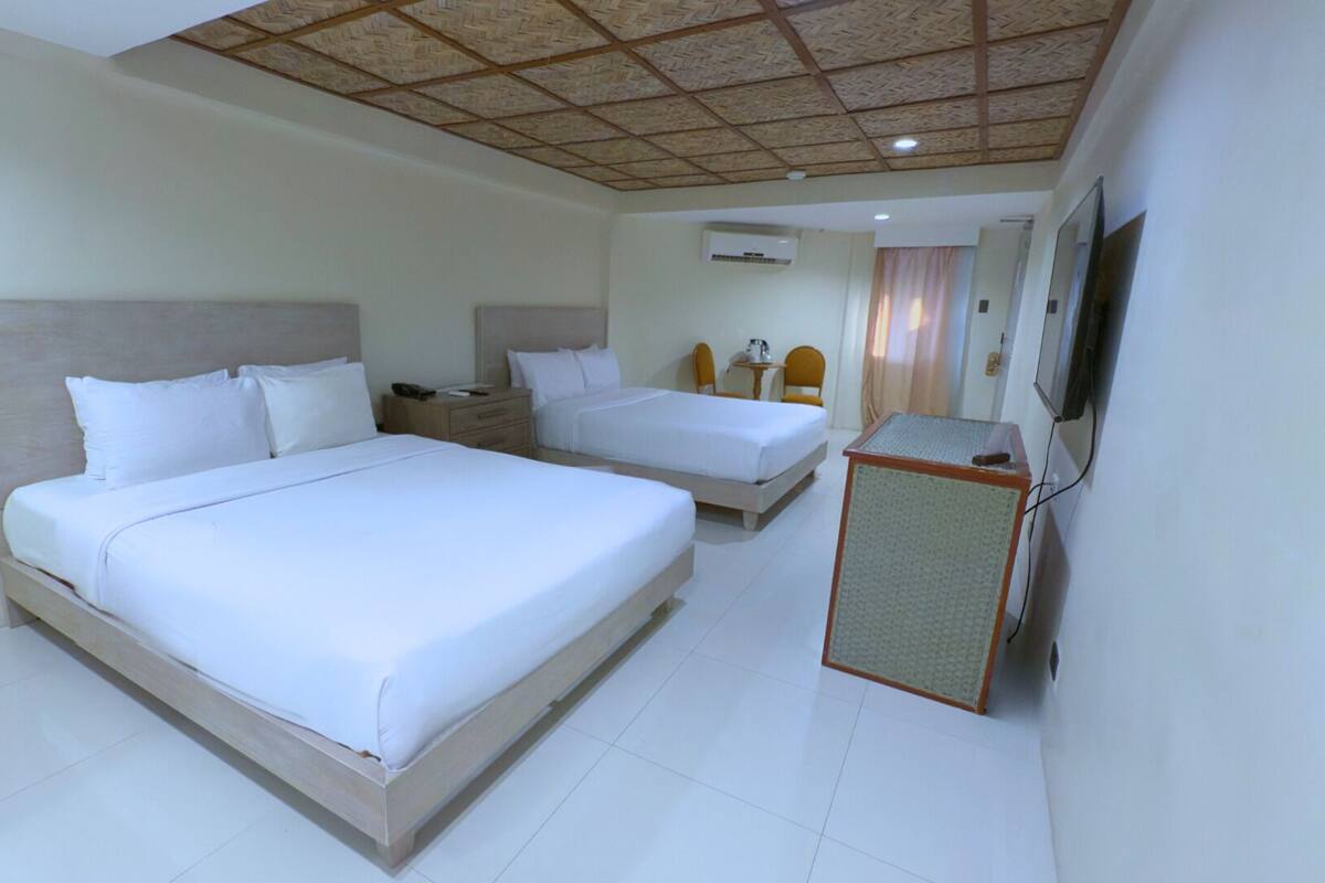 A hotel room with two beds, a small desk, a television, and an air conditioning unit.