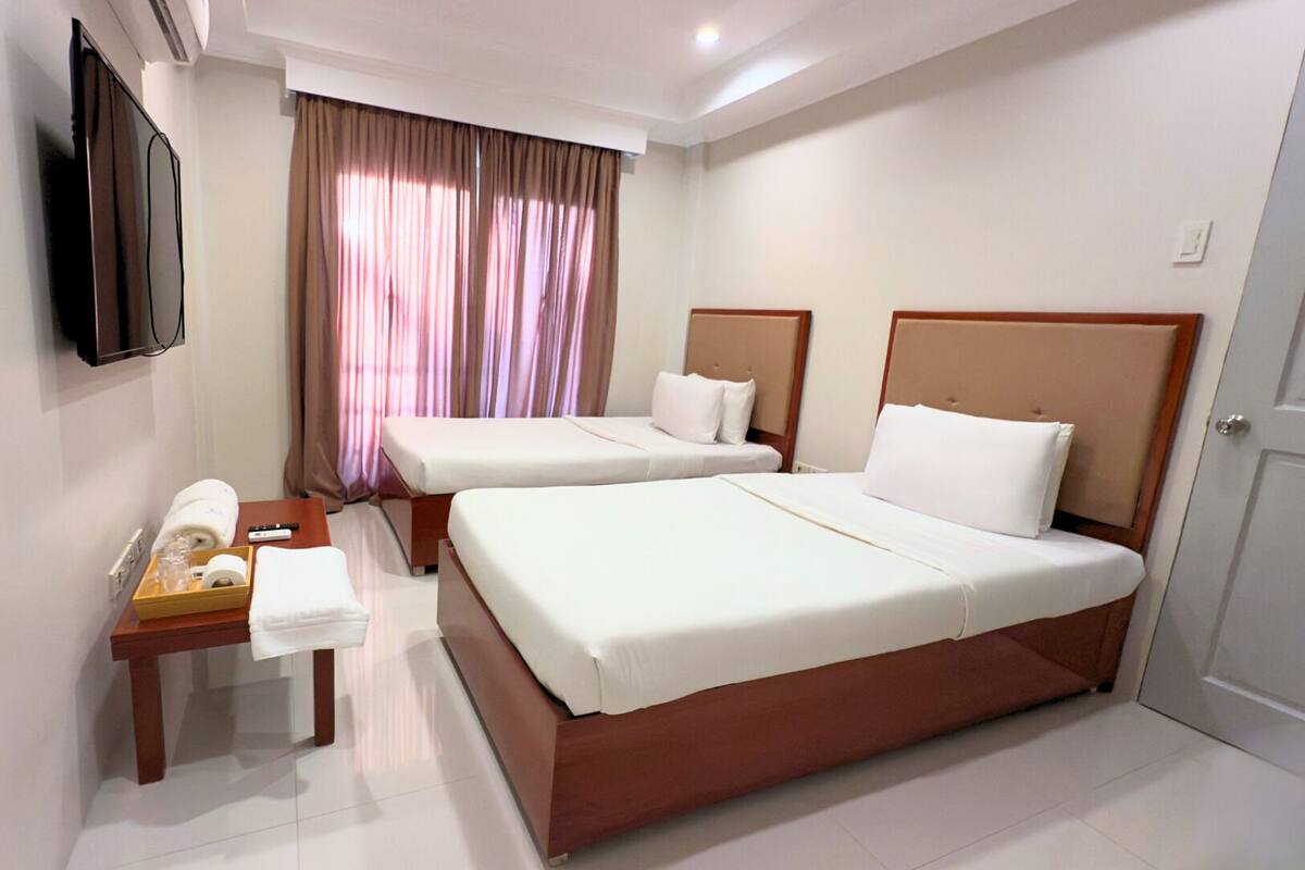 A hotel room with two beds, a TV, a bedside table with a lamp, and a window with curtains.