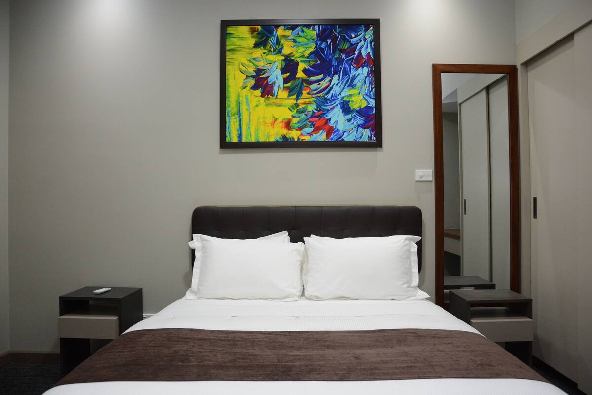 Habitación de hotel con una cama grande, dos mesitas de noche, un tocador y un cuadro en la pared, además de un espejo.