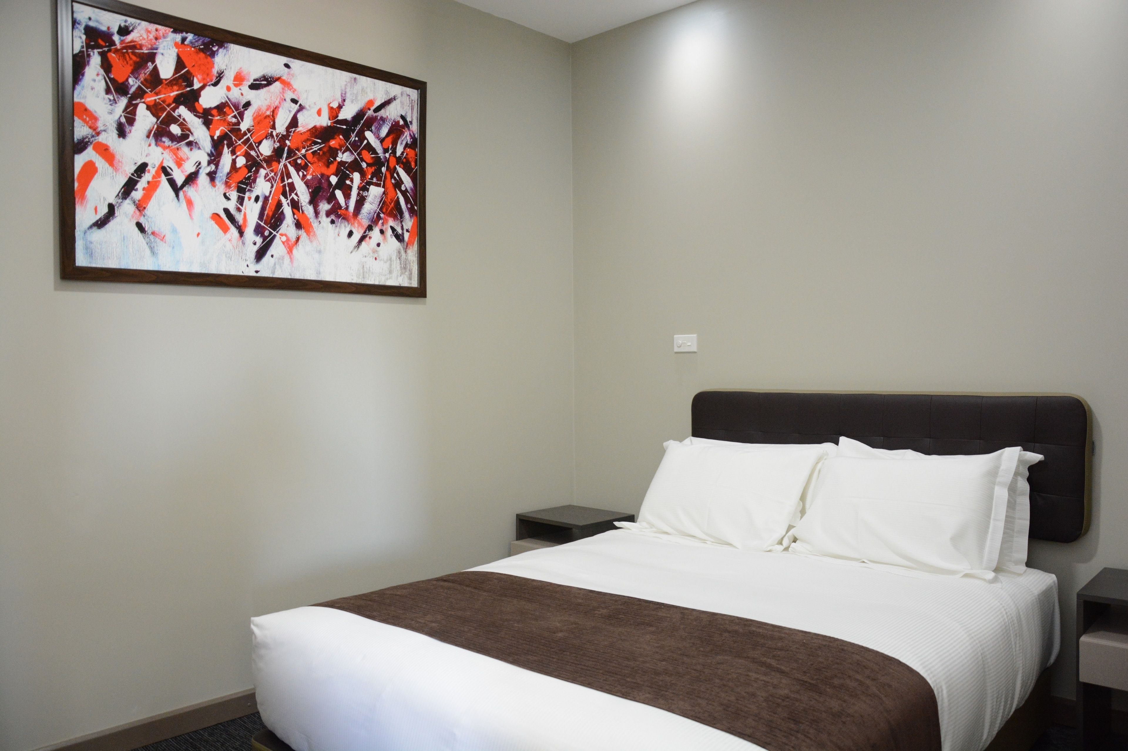 A hotel room with a large bed, a nightstand, a wall-mounted abstract painting, and a plain wall.