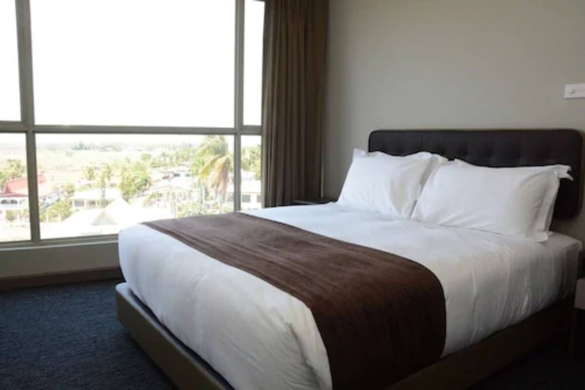 Una habitación de hotel con un ventanal grande, una cama con sábanas blancas y un plaid marrón.