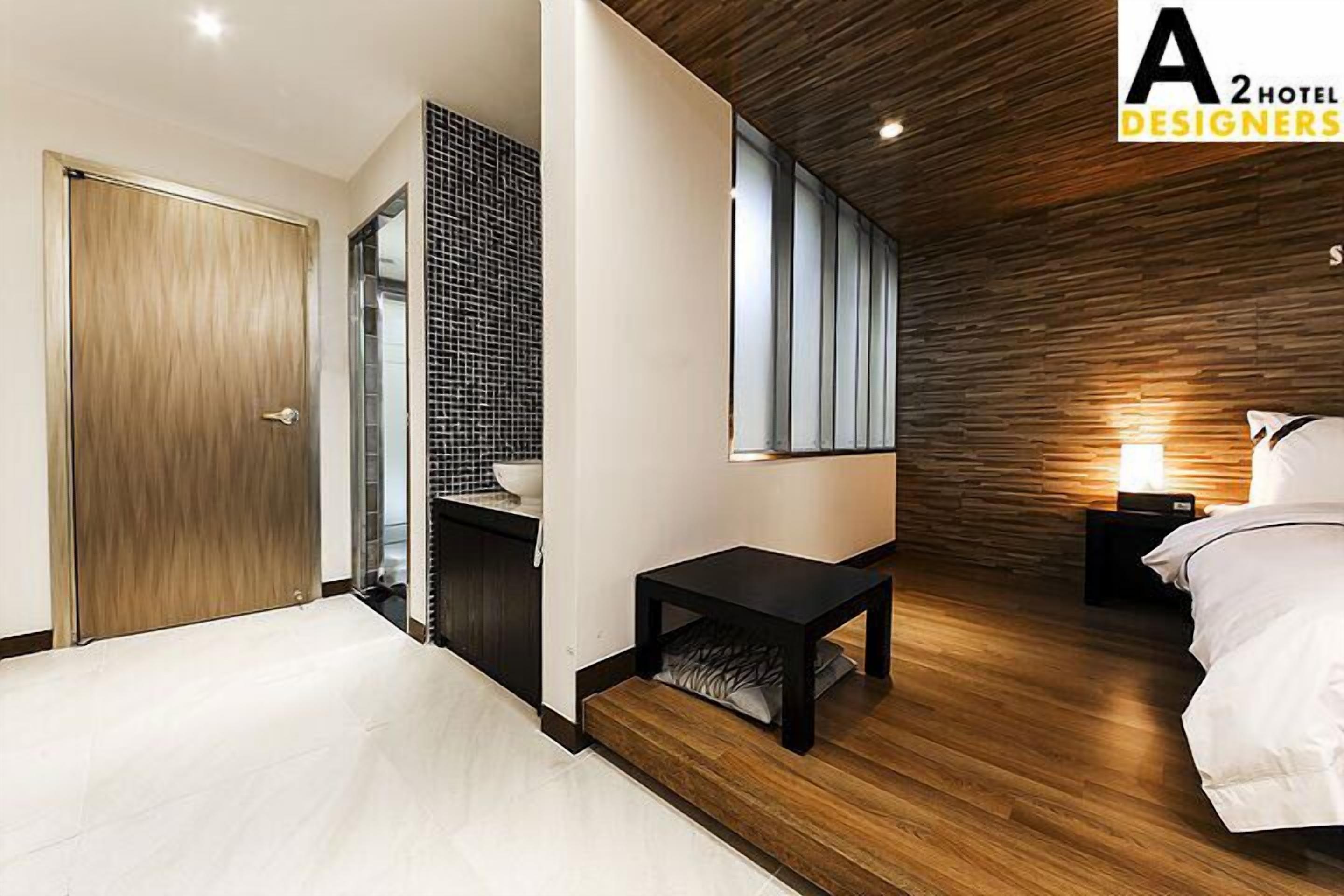 Una habitación de hotel moderna con piso de madera, una cama grande, una mesita pequeña, una mesita de noche y una lámpara fijada a la pared.