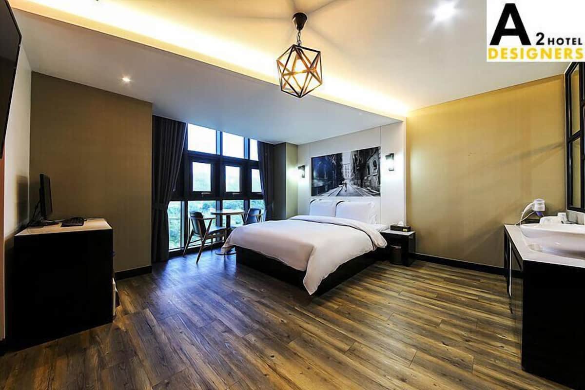 Una habitación de hotel moderna con piso de madera, una cama grande, un escritorio con una silla, una ventana con cortinas y un baño con lavamanos.