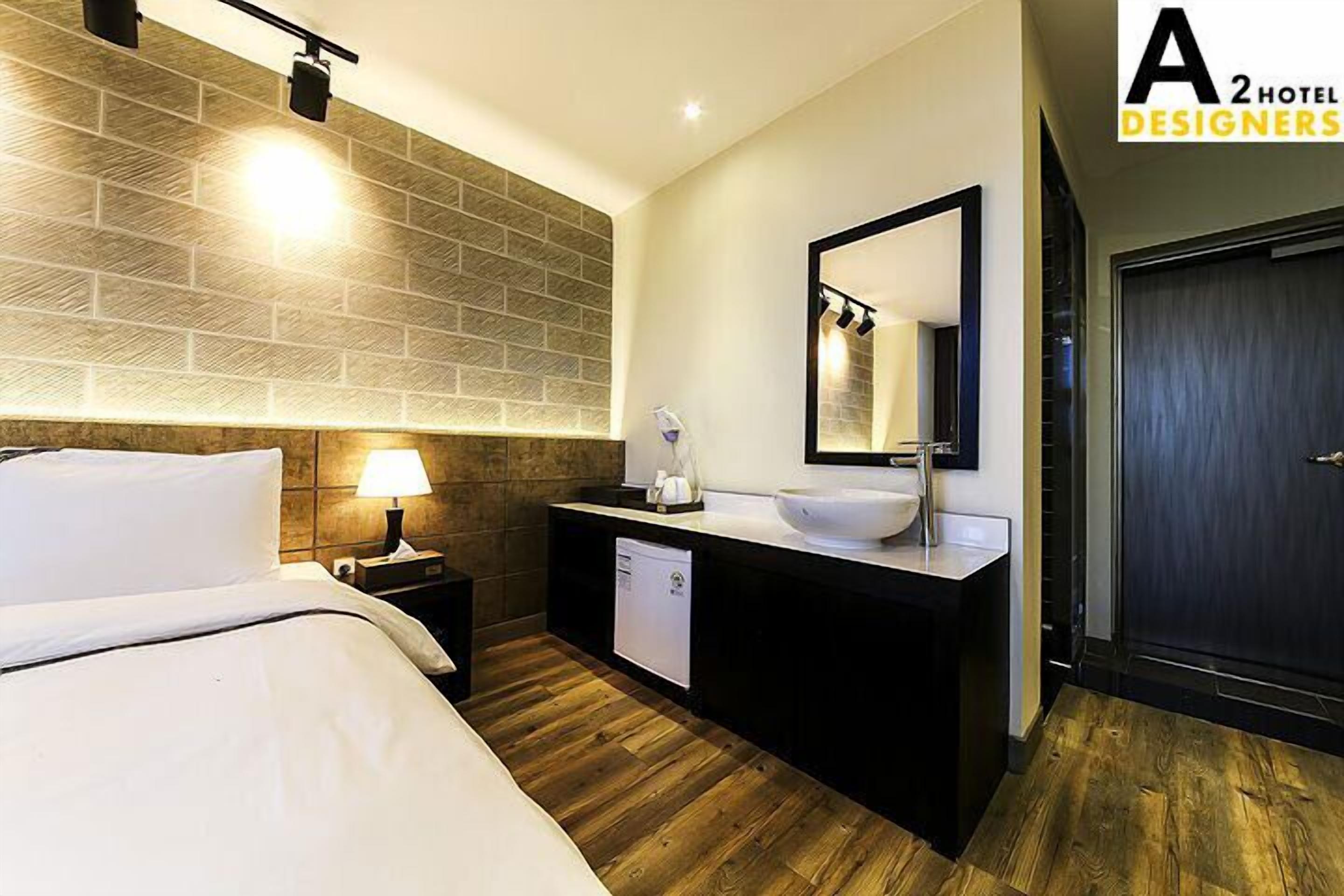 A hotel room with a bed, a bedside table with a lamp, a modern sink, and a dark door.