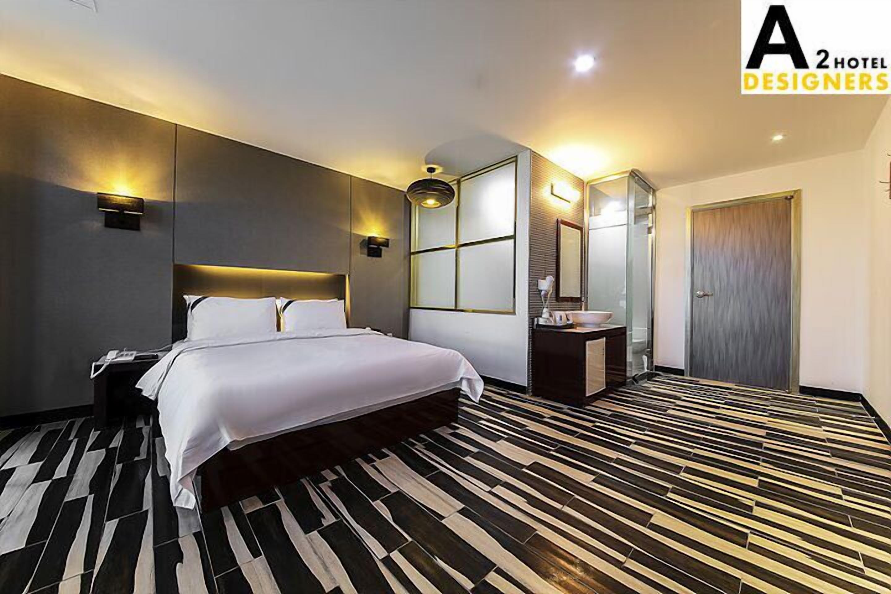 Ein modernes Hotelzimmer mit einem großen Bett, einem Nachttisch, einem Spiegel, einem kleinen Badebereich und einem gemusterten Boden.
