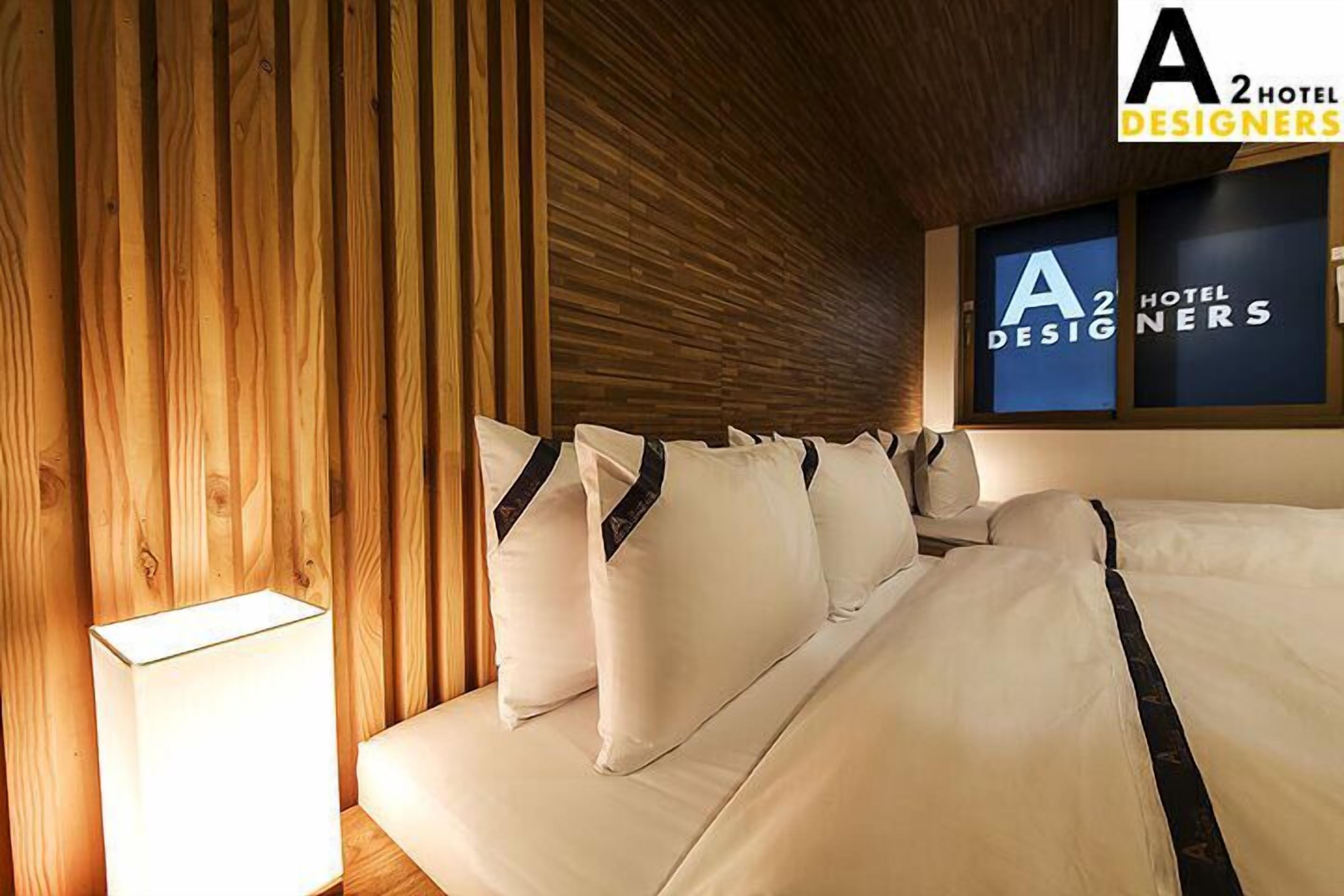 Habitación de hotel con pared de listones de madera, cama con ropa blanca y detalles oscuros, y letrero en la pared que dice 'A2 Hotel Designers'.