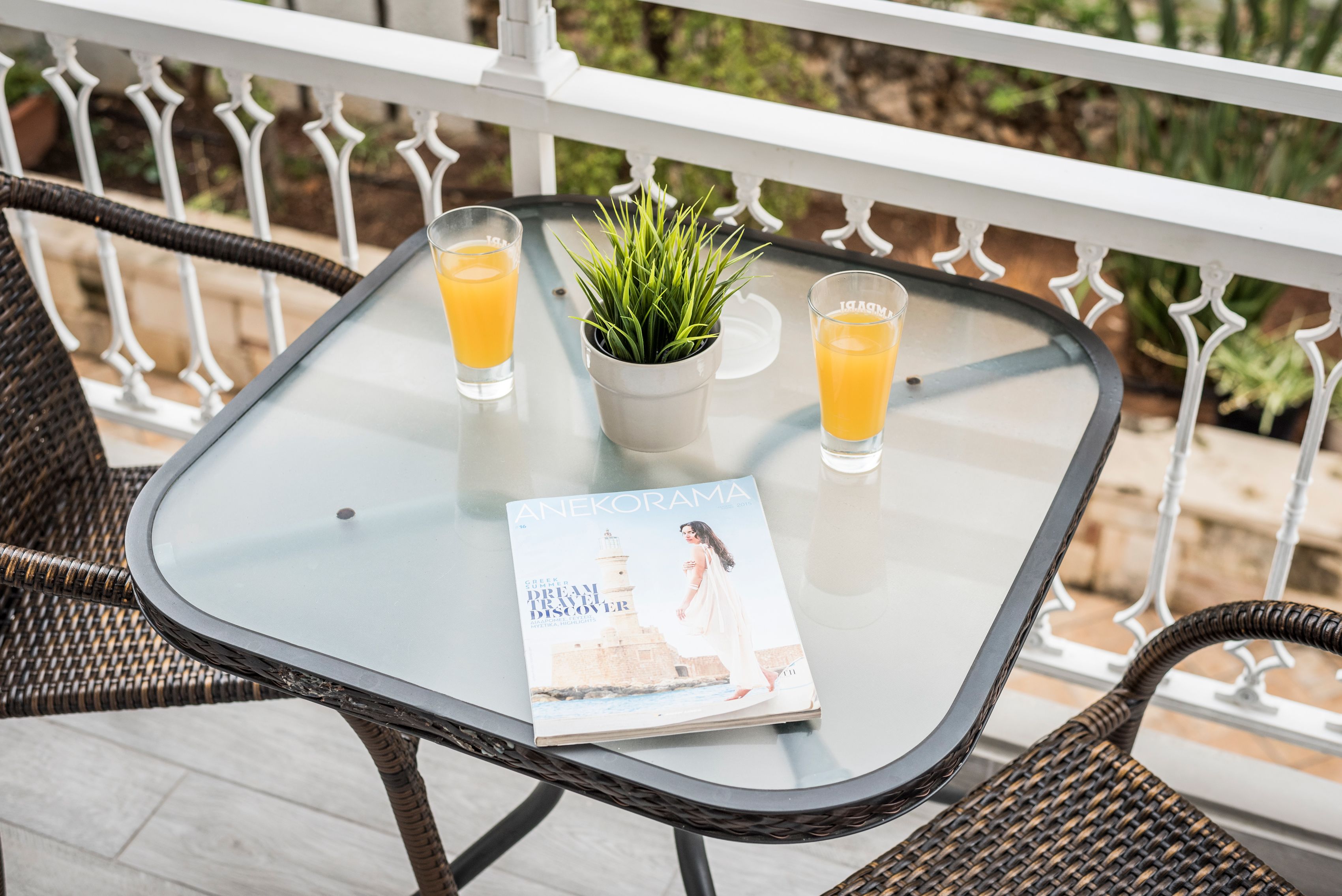 Ein Glastisch mit zwei Gläsern Orangensaft, einer Topfpflanze und einem Magazin mit dem Titel "Anekorama" auf einem Balkon mit Korbstühlen.