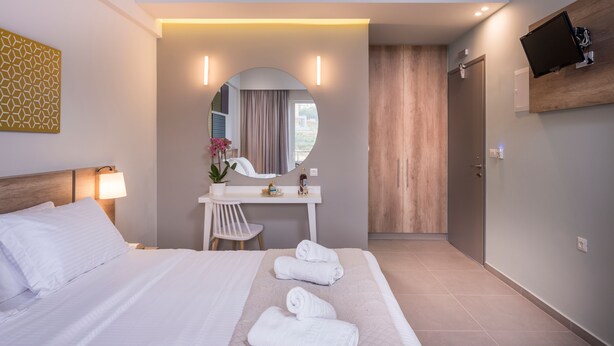 A modern hotel room with a bed, a desk, a mirror, and a TV mounted on the wall. - Irini Stalos Apartments (Chania)