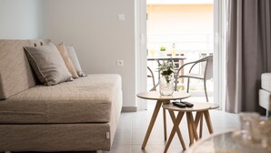 A modern living room with a beige sofa, a small round wooden table with a vase and remote control, and a view of an outdoor balcony. - Irini Stalos Apartments (Chania)