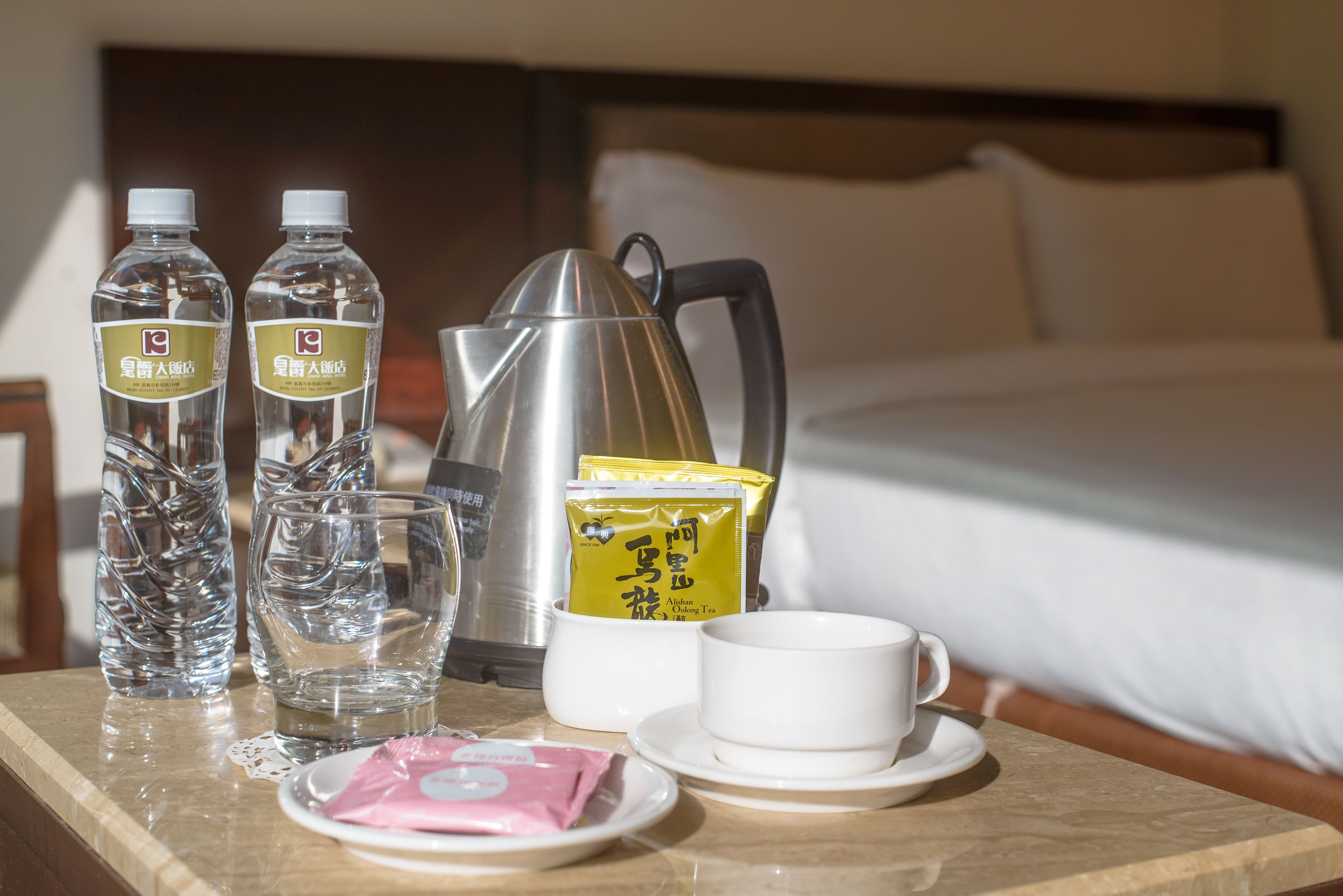 Een keurig opgemaakte hotelkamer met een bed, nachtkastje met waterkoker, twee waterflesjes, een beker en theezakjes.