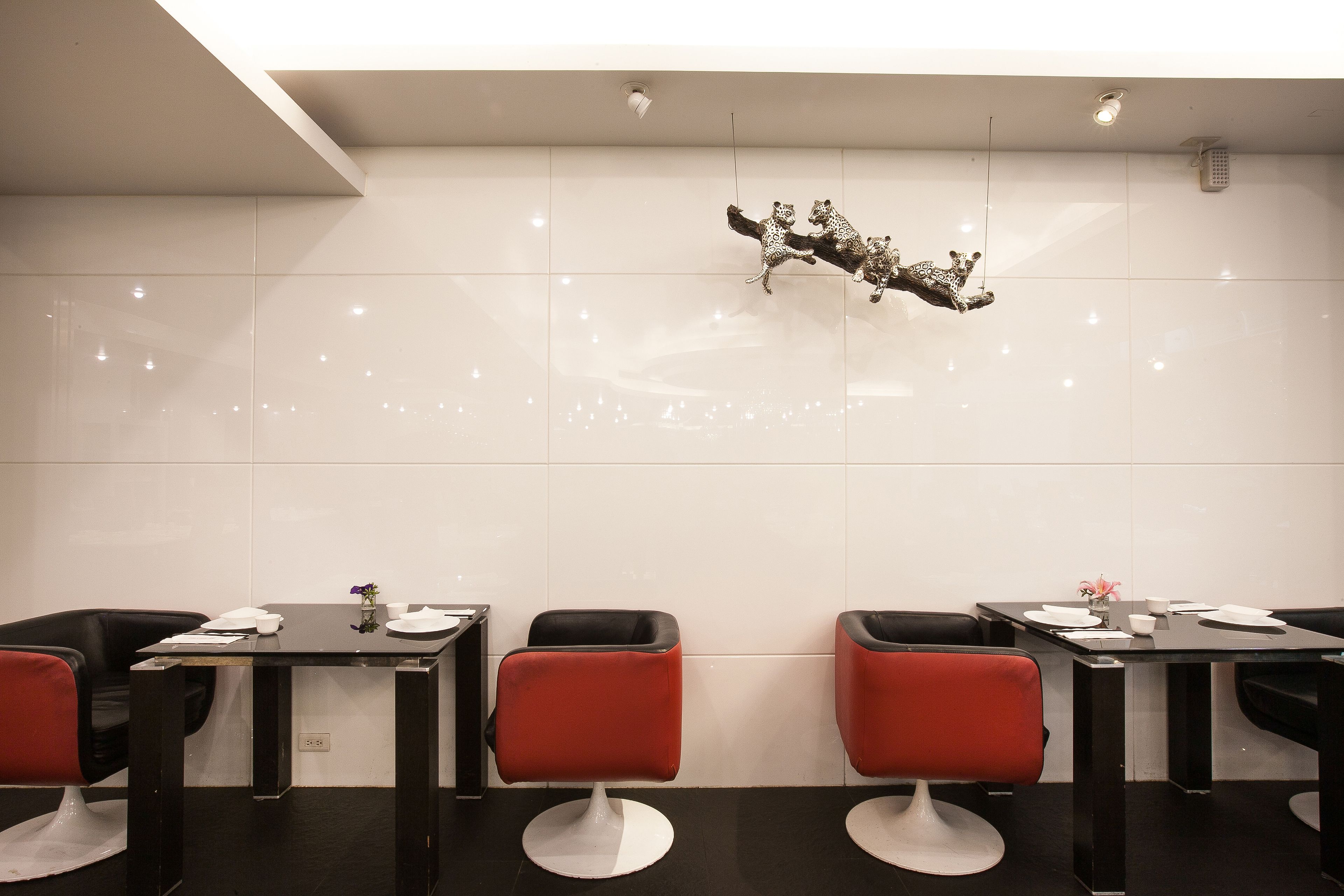 A modern restaurant with a clean, minimalist design, featuring black and red chairs, white-tiled walls, and a decorative metal sculpture.