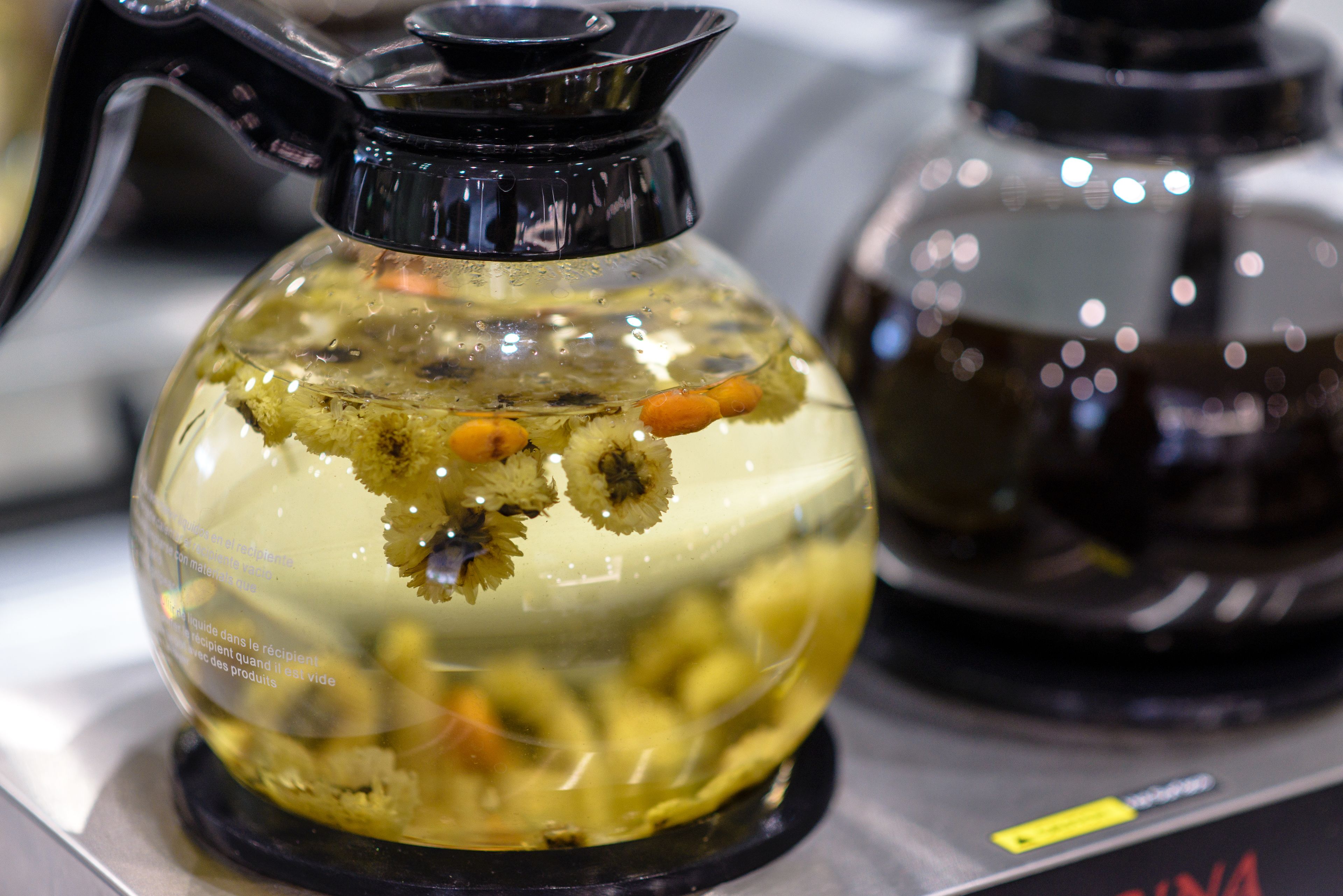 A clear glass container with a black spout, filled with a yellowish liquid and floating objects, possibly food items, on a reflective surface.